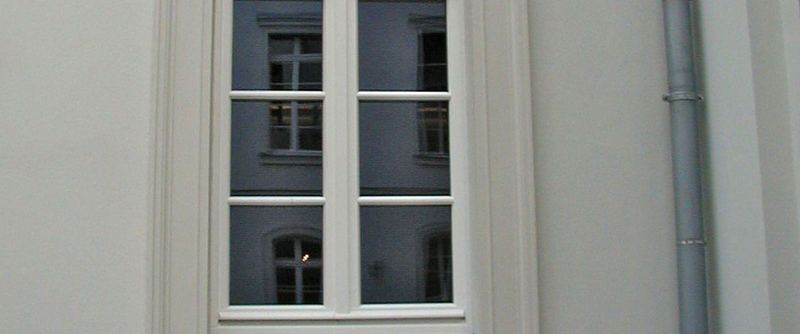 Zwei hohe Sprossenfenster in einer hellen Putzfassade, daneben ein Fallrohr.