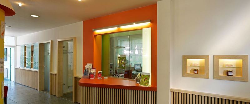 Empfangstresen mit orangefarbener Rückwand in einem hellen Eingangsbereich, daneben Türen und Wandverkleidung aus Holz.
