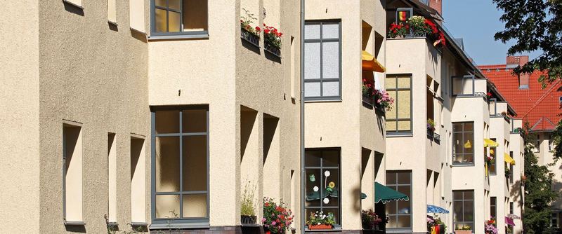 Straßenansicht eines mehrgeschossigen Wohnhauses mit hellen Holzfenstern, Blumenkästen und Markisen.